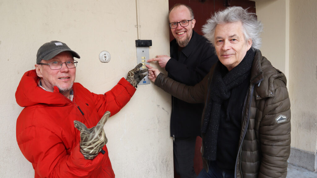 Kolme miestä talvitakeissa ulkona, osoittavat sormella nappia jossa numero 2. Kaikki katsovat kameraan.