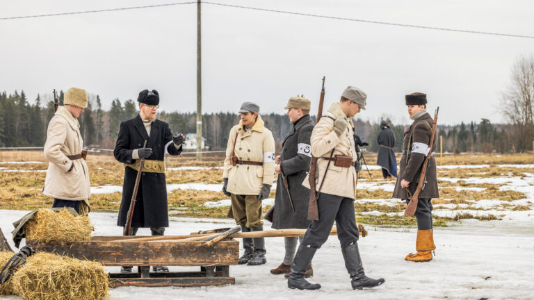 Miehiä talvitakeissa ja hatuissa. Käsissä ja olalla aseita. Taustalla peltomaisemaa.